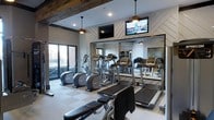 This well-appointed fitness center offers a thoughtfully designed workout environment that blends contemporary style with functional amenities. The space is anchored by a row of cardio machines, including treadmills and ellipticals, positioned in front of a full-length mirrored wall that enhances the sense of depth and openness throughout the room. A striking decorative wood beam runs across the ceiling, adding a warm, rustic contrast to the otherwise modern aesthetic. The walls feature a stylish chevron-patterned shiplap accent, contributing a clean yet textured visual element to the space. Overhead, pendant light fixtures provide warm ambient lighting, complemented by recessed ceiling lights for even illumination across the workout area. Multiple flat-screen televisions are mounted on the wall, ensuring entertainment is accessible during workouts. Large floor-to-ceiling windows along one side of the room invite abundant natural light and offer views of the surrounding exterior, creating an energizing atmosphere. The flooring consists of a checkered tile pattern that is both practical and visually appealing. Overall, this fitness center combines modern design details with a comprehensive selection of equipment, delivering a polished and motivating environment for exercise.