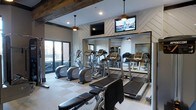 This well-appointed fitness center offers a thoughtfully designed workout environment that blends contemporary style with functional amenities. The space is anchored by a row of cardio machines, including treadmills and ellipticals, positioned in front of a full-length mirrored wall that enhances the sense of depth and openness throughout the room. A striking decorative wood beam runs across the ceiling, adding a warm, rustic contrast to the otherwise modern aesthetic. The walls feature a stylish chevron-patterned shiplap accent, contributing a clean yet textured visual element to the space. Overhead, pendant light fixtures provide warm ambient lighting, complemented by recessed ceiling lights for even illumination across the workout area. Multiple flat-screen televisions are mounted on the wall, ensuring entertainment is accessible during workouts. Large floor-to-ceiling windows along one side of the room invite abundant natural light and offer views of the surrounding exterior, creating an energizing atmosphere. The flooring consists of a checkered tile pattern that is both practical and visually appealing. Overall, this fitness center combines modern design details with a comprehensive selection of equipment, delivering a polished and motivating environment for exercise.