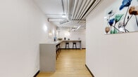 Located at 127 Northwest 27th Street in Miami's vibrant Wynwood neighborhood, this single-floor commercial suite offers a thoughtfully designed open-plan workspace with a contemporary industrial aesthetic. The space features light hardwood flooring throughout, crisp white walls, and exposed ceilings with visible ductwork and piping — a hallmark of modern office design that adds character and visual depth.

A standout feature is the glass-enclosed conference room, which provides a private meeting area while maintaining visual connectivity with the rest of the suite. Inside, a round table with mixed seating and a wall-mounted display screen make it well-suited for presentations and collaborative sessions. Adjacent to the conference room, a dedicated kitchenette area is equipped with cabinetry, a coffee station, and a compact refrigerator, supporting everyday workplace needs.

The open lounge area is furnished with high-back privacy chairs paired with side tables and anchored by a marble-patterned area rug, offering a comfortable setting for informal meetings or focused work. A long counter-height bar with bar stools runs along one wall, providing additional flexible workspace. Decorative touches — including a bold gold wall clock, curated artwork, and a large tropical potted plant — contribute to a polished, welcoming ambiance throughout the suite.