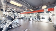 Located in Geneva, IL, this fitness center offers a comprehensive, single-floor workout facility designed to accommodate a wide range of exercise needs. The space features an industrial-style aesthetic with exposed ductwork, a dark painted ceiling, and large panel windows along multiple walls that flood the interior with natural light and provide views of the surrounding outdoor environment.

The main exercise area is divided into distinct training zones. The cardio section is equipped with multiple treadmills, elliptical trainers, stationary bikes, and a recumbent bike, all positioned along windowed walls to maximize natural light during workouts. A dedicated strength training zone features power racks, weight benches, and a full dumbbell rack, all reflected in a wall-to-wall mirror accented by a bold red horizontal stripe, adding a dynamic visual element to the space.

A functional training area provides open floor space alongside kettlebells, resistance bands, a rowing machine, and additional free weights, supported by a wall-mounted television for entertainment or instructional use.

The adjacent entry and lounge area features a welcoming seating space with a leather sofa, decorative plants, and large glass entry doors. A dedicated locker room area offers wood-toned lockers, coat hooks, bench seating, and restroom access, providing convenient amenities for users before and after their workouts. Durable rubber flooring throughout ensures safety and comfort across all areas of the facility.
