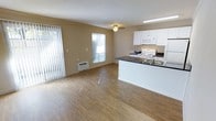 This single-floor space offers an open-concept layout that seamlessly integrates the living area and kitchen, creating a functional and inviting environment. The living area features warm hardwood-style flooring that extends throughout, contributing to a bright and cohesive aesthetic. Natural light filters in through multiple windows, including a large sliding glass door fitted with vertical blinds, enhancing the sense of openness. A ceiling fan provides additional comfort and airflow. A wall-mounted air conditioning unit ensures climate control within the living space. The kitchen is thoughtfully designed with white cabinetry, a dark countertop, and a peninsula that doubles as a divider between the cooking and living zones. White appliances, including a range, built-in microwave, and refrigerator, are neatly integrated into the cabinetry. A sink is conveniently located within the peninsula, making the kitchen both practical and accessible. The neutral beige wall tones throughout complement the flooring and cabinetry, giving the space a clean, understated look. Overall, the layout maximizes usability while maintaining an open and airy feel.