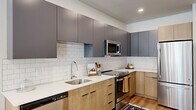 This single-floor, three-bedroom, two-bathroom residence offers a thoughtfully designed layout that balances functionality and modern aesthetics. The open kitchen is a standout feature, showcasing a two-tone cabinet design with slate-blue upper cabinets and natural light wood lower cabinets, complemented by a classic white subway tile backsplash and crisp quartz countertops. Stainless steel appliances, including a refrigerator, range, built-in microwave, and dishwasher, complete the space, while a central island with seating provides an ideal spot for casual dining. Light hardwood flooring flows throughout the unit, tying the spaces together cohesively. The living area is bright and welcoming, featuring recessed lighting and French doors that open to a private outdoor area, allowing natural light to flood in. Each bedroom is finished in neutral tones with ample natural light from wide windows fitted with blinds. The primary bedroom comfortably accommodates a full-size bed with room to spare. Both bathrooms are finished with light wood vanities, white quartz counters, and tub-shower combinations. The hallways are clean and bright, connecting each room efficiently, giving the home an organized, purposeful flow throughout.