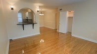 This single-story space showcases warm, polished hardwood flooring that extends throughout the open living and dining areas, reflecting light and adding a sense of spaciousness. A defining architectural feature is the elegant arched pass-through window connecting the kitchen to the living area, topped with a dark countertop breakfast bar supported by a decorative bracket — ideal for casual dining or entertaining. The kitchen, visible through the arch, features cabinetry and dark stone countertops with a tile backsplash.

The dining area is anchored by a traditional chandelier that adds a refined touch to the space. Neutral beige walls are complemented by white chair rail molding, lending a classic, well-appointed character to the interior. A visible HVAC vent is integrated into the wall, ensuring climate comfort throughout.

An open doorway leads to what appears to be a spacious closet or utility area with built-in shelving, adding practical storage to the layout. White trim and baseboards throughout tie the design together cohesively. The overall ambiance is bright, clean, and inviting, with traditional details balanced by an open, functional floor plan.