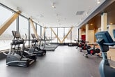 Located at 814 2e Avenue in Montréal, this well-appointed fitness facility offers a thoughtfully designed, single-level space spanning a generous footprint. The gym is defined by its striking floor-to-ceiling windows along one wall, fitted with distinctive diagonal wood-beam accents that serve as both a structural feature and a bold design statement. These large windows flood the interior with natural light and provide open views of the surrounding neighbourhood, creating an energizing atmosphere for workouts.

The cardio zone is equipped with a comprehensive lineup of True-brand treadmills, ellipticals, and stair climbers, all positioned to take advantage of the natural light and outdoor views. Nearby, a dedicated cycling area houses a row of spin bikes on durable rubber flooring. The strength and functional training zone features weight racks, benches, a cable machine, free weights, and an assortment of stability balls and medicine balls, supported by full-length mirrors along warm wood-panelled walls.

A separate movement and yoga zone offers light wood flooring and a darker, more intimate ceiling treatment, providing a calm contrast to the main gym. Recessed lighting and white structural columns frame this versatile space. Two bathrooms complete the facility, ensuring practical amenities for all users. The overall design balances modern aesthetics with functional fitness infrastructure.