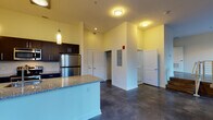 This single-floor unit showcases a contemporary open-concept design anchored by polished concrete floors that extend throughout the living and kitchen areas, lending an industrial-modern aesthetic. The kitchen is a standout feature, centered around a peninsula with granite countertops, an undermount sink, and dark espresso cabinetry. Stainless steel appliances—including a refrigerator, range, and built-in microwave—complete the kitchen, while sleek pendant lights hang above the peninsula, providing focused task lighting. The open living area benefits from a large window offering views of the surrounding urban streetscape, allowing natural light to fill the space. Elevated steps with a metal railing create a subtle separation within the open floor plan, adding architectural interest. The layout includes multiple doors leading to additional rooms and closet storage, with one area revealing a dedicated closet space. The bathroom features full tile surround walls in a neutral stone finish, a soaking tub with an overhead shower, and dark cabinetry. Throughout the unit, tall ceilings and a neutral color palette enhance the sense of openness, creating a clean and functional living environment.