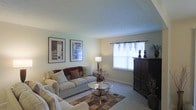 This single-floor space showcases a comfortable and thoughtfully arranged living area. The room is finished with soft, neutral-toned walls that create a calm and inviting atmosphere. Wall-to-wall carpeting in a warm beige adds to the sense of coziness underfoot, complemented by a patterned area rug that anchors the seating arrangement.

The living area is centered around a pair of plush, light-colored sofas accented with burgundy and cream throw pillows, offering ample seating. A glass-top coffee table with a decorative lower shelf serves as a functional centerpiece, while a matching glass side table holds a classic table lamp that provides warm ambient lighting.

A large window dressed with delicate sheer curtains allows natural light to filter into the room, brightening the space without sacrificing privacy. A dark wood entertainment armoire stands prominently against one wall, providing both storage and display space, with decorative candlestick accents on top.

Two framed art pieces adorn the walls, adding a polished, finished look to the overall décor. The space balances practicality with a welcoming, traditional aesthetic.