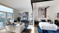Located at 1810 State Street in San Diego, this penthouse model unit — Plan K — is a single-floor residence that demonstrates a modern, open-concept design with carefully curated living spaces. The layout integrates the living and sleeping areas in a fluid arrangement, separated by a sleek sliding glass partition that allows natural light to pass freely throughout while still offering the option for privacy.

The living area features light hardwood flooring, white walls, and an abundance of natural light streaming in through large windows. A comfortable seating arrangement anchors the space, complemented by a dark area rug and a glass coffee table. The walls are adorned with an eclectic gallery of framed artwork, adding personality and visual interest to the room. A media console with a mounted television serves as a functional focal point.

The sleeping area is distinguished by a dramatic floor-to-ceiling architectural photographic mural, lending an artistic and cosmopolitan character to the room. A well-appointed bed with layered bedding sits atop a deep navy rug, creating a cozy, defined retreat. The overall ambiance throughout the unit is contemporary and refined, blending urban sophistication with comfortable, livable design.