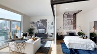 Located at 1810 State Street in San Diego, this penthouse model unit — Plan K — is a single-floor residence that demonstrates a modern, open-concept design with carefully curated living spaces. The layout integrates the living and sleeping areas in a fluid arrangement, separated by a sleek sliding glass partition that allows natural light to pass freely throughout while still offering the option for privacy.

The living area features light hardwood flooring, white walls, and an abundance of natural light streaming in through large windows. A comfortable seating arrangement anchors the space, complemented by a dark area rug and a glass coffee table. The walls are adorned with an eclectic gallery of framed artwork, adding personality and visual interest to the room. A media console with a mounted television serves as a functional focal point.

The sleeping area is distinguished by a dramatic floor-to-ceiling architectural photographic mural, lending an artistic and cosmopolitan character to the room. A well-appointed bed with layered bedding sits atop a deep navy rug, creating a cozy, defined retreat. The overall ambiance throughout the unit is contemporary and refined, blending urban sophistication with comfortable, livable design.