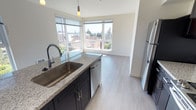 Located on the fifth floor at 4505 42nd Ave SW in Seattle's West Seattle neighborhood, this modern unit offers a thoughtfully designed single-level layout filled with natural light. The open-concept kitchen is a standout feature, anchored by a generous granite-topped island with an undermount sink and pendant lighting overhead. Dark espresso cabinetry contrasts elegantly with the light granite countertops, while stainless steel appliances — including a refrigerator, dishwasher, microwave, and range — complete the well-equipped space. Corner windows in the kitchen and living area frame sweeping views of the surrounding neighborhood and green treetops, flooding the space with daylight. The bedrooms feature light wood-tone or carpeted flooring and large windows that continue the bright, airy feel. One bedroom includes frosted glass sliding closet doors, adding a sleek, contemporary touch. The bathroom is finished with matching granite countertops, dark cabinetry, a wide vanity mirror, and durable wood-tone flooring consistent with the rest of the unit. Throughout, the clean white walls, modern finishes, and cohesive design palette create a polished and move-in-ready living environment in one of Seattle's most sought-after neighborhoods.