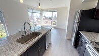 Located on the fifth floor at 4505 42nd Ave SW in Seattle's West Seattle neighborhood, this modern unit offers a thoughtfully designed single-level layout filled with natural light. The open-concept kitchen is a standout feature, anchored by a generous granite-topped island with an undermount sink and pendant lighting overhead. Dark espresso cabinetry contrasts elegantly with the light granite countertops, while stainless steel appliances — including a refrigerator, dishwasher, microwave, and range — complete the well-equipped space. Corner windows in the kitchen and living area frame sweeping views of the surrounding neighborhood and green treetops, flooding the space with daylight. The bedrooms feature light wood-tone or carpeted flooring and large windows that continue the bright, airy feel. One bedroom includes frosted glass sliding closet doors, adding a sleek, contemporary touch. The bathroom is finished with matching granite countertops, dark cabinetry, a wide vanity mirror, and durable wood-tone flooring consistent with the rest of the unit. Throughout, the clean white walls, modern finishes, and cohesive design palette create a polished and move-in-ready living environment in one of Seattle's most sought-after neighborhoods.