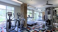 This fitness center is a thoughtfully designed workout space that combines functional amenities with distinctive architectural character. The room is anchored by striking exposed brick columns that add an industrial aesthetic and visual interest throughout the space. Large windows line the walls, flooding the interior with abundant natural light and offering views of the surrounding greenery, creating an energizing atmosphere for exercise.

The facility is well-stocked with cardio equipment, including multiple treadmills and elliptical machines, arranged to take full advantage of the natural light and outdoor views. A floor-to-ceiling mirror along one wall expands the sense of space while allowing members to monitor their form during workouts. Ceiling fans and pendant lighting fixtures provide both comfort and even illumination throughout.

Underfoot, a vibrant geometric-patterned carpet in bold reds, oranges, greens, and whites adds a lively, modern touch to the space. The overall design balances classic brick detailing with contemporary finishes, resulting in a welcoming and motivating environment. The single-level layout ensures easy accessibility, and the open floor plan provides ample room to move freely between equipment.