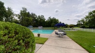 This single-level outdoor facility is centered around a generously sized rectangular swimming pool with clear, turquoise water, making it an inviting focal point of the property. The pool deck is finished in smooth concrete and lined with numerous lounge chairs positioned for optimal sun exposure, along with seating areas featuring white tables and chairs shaded by navy blue umbrellas — ideal for relaxing or socializing. A dedicated storage shed with double doors sits adjacent to the pool area, providing convenient access to equipment and supplies. The pool is enclosed by a metal fence that ensures safety while preserving open sightlines to the surrounding landscape.

The grounds are beautifully maintained, with lush green lawns, mature trees forming a natural backdrop, and neatly trimmed shrubs bordering the brick building exterior. The building itself is constructed of classic red brick with white trim, contributing a timeless and well-kept aesthetic. A covered entryway leads into the interior, featuring a distinctive blue paneled door with glass inserts, offering a welcoming transition between the outdoor and indoor spaces. Concrete walkways connect the various areas of the property, providing easy and accessible circulation throughout. The overall ambiance is one of a well-appointed, community-oriented recreational facility set within a verdant, park-like setting.