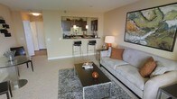 This single-floor space boasts an open-concept layout that seamlessly integrates the living area and kitchen, creating a cohesive and functional environment. The living area features plush carpeting, a contemporary sofa with accent pillows, and a sleek rectangular coffee table set atop a textured area rug, contributing to a warm and inviting atmosphere. A framed abstract artwork adorns the wall, adding a touch of modern sophistication. The kitchen is elevated behind a raised breakfast bar with two stylish bar stools, providing both a casual dining option and a natural visual division between spaces. Dark espresso cabinetry contrasts elegantly with lighter walls and countertops, while pendant lighting above the bar adds a refined finishing touch. Stainless steel appliances, including a microwave and refrigerator, are visible within the kitchen, underscoring the space's modern sensibility. Neutral, warm-toned walls throughout unify the space and complement the overall contemporary aesthetic. The open flow between areas enhances the sense of spaciousness, making this layout ideal for both everyday living and casual entertaining.