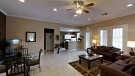 This single-story space offers an open, well-connected layout that blends functional and comfortable areas seamlessly. The main living or lounge area is anchored by plush dark upholstered sofas arranged around a wooden coffee table atop a striped area rug, creating a warm and inviting atmosphere. A ceiling fan with a light fixture overhead provides both airflow and ambient lighting, complementing the recessed lighting throughout. Crown molding along the upper walls adds a refined architectural touch.

Adjacent to the seating area is a small dining nook, furnished with a glass-top table and elegant chairs, offering an intimate setting. The space opens toward a counter or reception area, distinguished by white cabinetry and overhead fluorescent lighting, suggesting a service or check-in function. This area is flanked by a doorway leading to a separate corridor or back room, adding depth to the overall floor plan.

Natural light filters in through multiple windows and a glass-paneled door, brightening the light-toned ceramic tile flooring that runs throughout the space. Potted plants and decorative accents add warmth, while the neutral wall tones and white trim contribute to a clean, cohesive aesthetic overall.