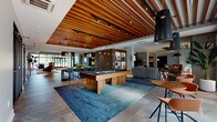 Located at 111 Route 70 East in Evesham, NJ, this single-floor amenity space is a masterfully designed communal lounge that blends contemporary aesthetics with a welcoming, social atmosphere. The space is anchored by striking slatted wood ceiling panels that run throughout, adding warmth and architectural character while complementing the track lighting above.

The lounge area features a large sectional sofa arranged around a linear gas fireplace set into a sleek concrete-panel surround, topped by a wall-mounted flat-screen TV — creating an inviting focal point for relaxation. Adjacent to the lounge sits a dedicated card and game table area, illuminated by a bold dome pendant light and framed by expansive windows overlooking an outdoor pool area, flooding the space with natural light.

A centrally positioned billiards table rests on a deep blue area rug, surrounded by tile flooring and high-top bar seating, serving as the entertainment hub of the room. The open-plan layout flows seamlessly between activity zones, including a bar-style dining counter and shelved display area with warm wood paneling.

A separate kitchen nook features stainless steel appliances, dark cabinetry, open shelving with a decorative tile backsplash, and a solid wood dining table — ideal for catered gatherings or casual use. Throughout, the design balances bold color accents with natural materials for a refined yet lively communal environment.