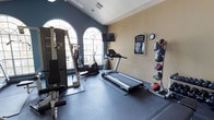 This fitness center offers a thoughtfully designed workout space that combines functionality with an inviting atmosphere. The room is anchored by striking arched windows that flood the space with natural light, creating an energizing environment for exercise. The walls are finished in a two-tone palette of cool slate blue and warm beige, lending the space a polished, professional appearance.

The flooring consists of durable dark rubber matting, providing both safety and comfort during workouts. Cardio equipment includes a treadmill and an elliptical machine, both positioned to take advantage of the natural light from the arched windows. A multi-station strength training machine is also present, offering a range of resistance exercises. A tiered medicine ball rack along the wall provides additional options for functional and core training.

A wall-mounted flat-screen television is positioned for easy viewing during workouts, while a clock keeps members on schedule. A "Fitness Center" sign with posted guidelines is displayed on the wall, contributing to an organized and informative environment. The vaulted ceiling adds a sense of openness and volume to the space, enhancing the overall sense of comfort. This fitness center is well-suited for a well-rounded exercise routine in a clean, bright, and welcoming setting.