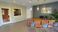 This single-story commercial space presents a polished and professional environment centered around a well-appointed reception area. The focal point is a warm, wood-toned reception desk with a textured finish, complemented by modern gray guest chairs that invite a comfortable client experience. Three elegant glass teardrop pendant lights hang overhead, casting a warm glow that contrasts beautifully with the recessed ceiling lighting throughout.

The space features a two-tone color scheme, pairing soft gray walls with a bold, darker charcoal accent wall behind the reception desk, where a branded logo serves as a visual centerpiece. A large interior window on the adjacent wall offers a view into a secondary area, adding depth and transparency to the layout while maintaining a sense of openness.

The flooring consists of neutral-toned large-format tiles, contributing to the clean and contemporary feel of the space. A white interior door provides access to additional rooms beyond the reception area. Overall, the space conveys a professional, welcoming atmosphere well-suited for client-facing business operations.