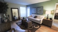 This single-floor residence offers a comfortable and stylishly appointed living space designed with both function and aesthetic appeal in mind. The living room serves as the heart of the home, showcasing warm hardwood-style flooring that runs throughout and creates a cohesive, inviting atmosphere. A bold teal accent wall provides a striking focal point, complemented by sage green walls that add depth and a calming, nature-inspired palette to the room. Two framed nature-themed art prints adorn the accent wall, enhancing the space's relaxed yet polished character. Natural light filters in through a window fitted with classic white blinds, brightening the interior and creating an airy feel. The living area is well-proportioned, offering ample room for comfortable seating arrangements, as evidenced by the cream sofa, woven ottoman coffee tables, and a patterned area rug that anchor the space. A large floor mirror along one wall amplifies the sense of space and reflects additional light throughout the room. The textured ceiling adds a subtle traditional detail overhead. Overall, the residence presents a warm, well-balanced environment that blends comfortable living with thoughtful design touches throughout.