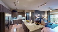 This thoughtfully designed two-bedroom, two-bathroom residence in Spring, Texas, offers a cohesive open-concept layout across a single floor. The kitchen serves as a central hub, showcasing granite-style countertops, a substantial island with pendant lighting, dark wood cabinetry, stainless steel appliances, a tile backsplash, and track lighting overhead. Wood-look flooring flows throughout the main living areas, adding warmth and continuity.

The living room features a ceiling fan, sliding glass doors that open to a private balcony, and large windows that invite natural light. The primary bedroom includes plush carpeting, a bold accent wall, and a window workspace area. Its en-suite bathroom offers a dual-sink vanity with granite countertops, dark cabinetry, a walk-in shower with glass enclosure, and a walk-in closet. The second bedroom is equally well-appointed, with natural light and a warm accent wall. The second bathroom provides a full tub-shower combination and matching vanity finishes. An in-unit washer and dryer area adds convenience, completing this well-equipped residence.
