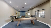 This single-floor recreational space is designed as a dedicated game room and social hub. The centerpiece of the room is a full-sized shuffleboard table with a natural wood finish, complemented by a pool table with a blue felt surface and a complete set of billiard balls. The room is finished with wall-to-wall blue-gray carpeting that provides a comfortable and cohesive aesthetic underfoot. Recessed lighting throughout the textured ceiling ensures the space is well-lit for gameplay and socializing. Bar-height tables with swivel stools are positioned around the perimeter, offering casual seating for spectators and guests. Colorful billiard-themed wall art adds a playful, thematic accent to the neutral beige and slate-blue painted walls. Large windows along one wall allow natural light to filter in and offer views of a landscaped outdoor area. A full-length mirror on one wall visually expands the space. The game room connects openly to adjacent areas, including a lounge with seating and a separate workspace or business center, making this a versatile and well-integrated amenity space suited for both recreation and relaxed social gatherings.
