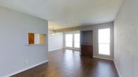 This single-floor space offers a welcoming and open layout that seamlessly connects the living and dining areas, creating a sense of spaciousness and ease of movement throughout. Rich, dark hardwood flooring runs continuously across the main living area, adding warmth and a polished aesthetic to the interior.

Natural light is a defining feature of this space, with multiple large windows along the exterior wall flooding the rooms with brightness and creating an airy, open ambiance. The neutral gray wall tones complement the flooring and allow the natural light to reflect evenly throughout.

A distinctive built-in cabinet niche is recessed into one wall, providing a dedicated space for storage or display and adding a thoughtful architectural detail to the living area. A pendant light fixture overhead in the dining area serves as a functional and decorative focal point, defining the dining zone within the open floor plan.

The kitchen connects to the main living area via a pass-through window framed by warm wood cabinetry, allowing for easy interaction between spaces while maintaining a degree of separation. White baseboards and clean wall finishes contribute to a crisp, well-maintained appearance throughout. Overall, the space combines practical design with inviting details to create a comfortable and functional single-level environment.