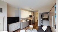 This thoughtfully designed studio unit maximizes functionality through a smart, open-concept layout. The living and kitchen areas share a unified space anchored by rich, dark hardwood flooring that runs throughout. The kitchen features a sleek galley-style configuration with white upper cabinetry, light wood-toned lower cabinets, a tiled backsplash, stainless steel appliances including a refrigerator, microwave, and dishwasher, and a single-basin sink with modern fixtures. Track lighting overhead illuminates both the kitchen and living areas efficiently.

The living space is highlighted by a built-in Murphy bed system flanked by integrated open shelving, offering versatile use as both a sleeping and living area. A gray sofa and a small workspace with a built-in desk complete the multifunctional layout. A large window allows natural light to enter, enhancing the open feel of the space.

Storage is addressed through a dedicated closet with built-in shelving, visible from the main living area. The bathroom is clean and minimal, featuring light wood-toned cabinetry, a white countertop vanity, light tile flooring, and neutral white walls, creating a calm, functional space.