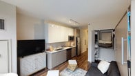 This thoughtfully designed studio unit maximizes functionality through a smart, open-concept layout. The living and kitchen areas share a unified space anchored by rich, dark hardwood flooring that runs throughout. The kitchen features a sleek galley-style configuration with white upper cabinetry, light wood-toned lower cabinets, a tiled backsplash, stainless steel appliances including a refrigerator, microwave, and dishwasher, and a single-basin sink with modern fixtures. Track lighting overhead illuminates both the kitchen and living areas efficiently.

The living space is highlighted by a built-in Murphy bed system flanked by integrated open shelving, offering versatile use as both a sleeping and living area. A gray sofa and a small workspace with a built-in desk complete the multifunctional layout. A large window allows natural light to enter, enhancing the open feel of the space.

Storage is addressed through a dedicated closet with built-in shelving, visible from the main living area. The bathroom is clean and minimal, featuring light wood-toned cabinetry, a white countertop vanity, light tile flooring, and neutral white walls, creating a calm, functional space.