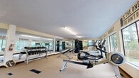 Located at 1923 Purcell Way in North Vancouver, BC, this fitness facility offers a comprehensive workout environment designed to support a wide range of exercise routines. The single-floor gym is generously sized, providing ample floor space for both equipment and open movement.

The main workout area is lined with floor-to-ceiling mirrors along one wall, enhancing the sense of space while allowing users to monitor their form. A well-curated selection of cardio equipment includes treadmills, an elliptical trainer, a stationary bike, and a rowing machine, all positioned to take advantage of the natural light streaming in through large windows that frame views of surrounding lush greenery. A dedicated free weights section features a full dumbbell rack with a weight bench, catering to strength training needs.

A separate open area at one end of the facility provides a dedicated space for stretching, floor exercises, and functional training, complete with exercise mats and a stability ball. A wall-mounted water fountain offers convenient hydration. Motivational phrases are displayed prominently on the walls, contributing to an energizing atmosphere. The textured ceiling, neutral carpet flooring, and bright fluorescent lighting complete the practical and focused ambiance of this well-appointed fitness space.