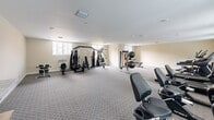 Located at 2203 N University Dr in Pembroke Pines, Florida, this fitness center offers a single, generously sized exercise room designed to accommodate a comprehensive range of workout activities. The space is finished with neutral, warm-toned walls and durable patterned carpet flooring, creating a clean and functional environment conducive to exercise.

The room is well-lit by multiple windows positioned along the walls, allowing ample natural light to flow in and providing views of the surrounding exterior. Recessed ceiling fixtures supplement the natural light, ensuring a bright and inviting atmosphere throughout.

The fitness center is equipped with a diverse selection of exercise machines and equipment. Cardio offerings include multiple treadmills and elliptical trainers, along with stationary cycling machines, catering to a variety of cardiovascular workout preferences. For strength training, the space features a multi-station cable and weight machine, free weights including medicine balls and dumbbells, resistance bands, and exercise stability balls. Additional benches and a rowing machine round out the strength and conditioning options.

Wall-mounted fans provide supplemental air circulation, and clearly marked emergency exits ensure safety. The open, spacious layout allows for comfortable movement between equipment, making this fitness center a practical and well-appointed facility for residents and users of the Pembroke Pines community.