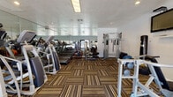Located at 1800 Stokes St in San Jose, California, this well-equipped fitness center offers a comprehensive range of exercise options within a single-floor layout. The space is finished with distinctive striped carpet tiles in earthy tones of brown, tan, and black, lending a modern and energetic aesthetic throughout.

The cardio zone is anchored by a row of treadmills and elliptical machines, neatly aligned along a full-length mirrored wall that visually expands the space and allows users to monitor their form during workouts. The mirrors also reflect natural light, contributing to a bright and open atmosphere.

Strength training is well represented with a multi-function cable machine, a dedicated weight rack stocked with dumbbells, a stability ball, and a freestanding punching bag — catering to a variety of training styles. A wall-mounted flat-screen television provides entertainment during workouts.

Large windows and a set of glass doors at the rear of the room look out directly onto an outdoor pool area, offering pleasant views and a strong connection to the exterior amenity space. Recessed and fluorescent lighting ensure the interior remains well-lit at all times. The textured ceiling and clean white walls complete the functional yet comfortable environment, making this fitness center a practical and inviting space for residents.