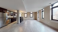 This two-bedroom, two-bathroom residence offers a generously sized, single-level floor plan designed with modern urban living in mind. The open-concept living and dining area is anchored by light-toned porcelain tile flooring and framed by multiple large windows with dark trim, including arched accents, that frame city skyline views and fill the space with natural light.

The kitchen is thoughtfully designed with rich espresso-stained cabinetry, stone countertops, stainless steel appliances including a range, microwave, refrigerator, and dishwasher, and a peninsula with pendant lighting overhead — ideal for both cooking and casual dining.

Both bedrooms are carpeted and well-proportioned, with large windows offering urban views and ample wall space for furnishings. The primary bathroom features a glass-enclosed walk-in shower, dark vanity cabinetry, stone countertops, and tile flooring with a mosaic border accent. The second bathroom includes a tub-shower combination, matching dark cabinetry, and coordinating tile work. A hallway with built-in shelving provides additional storage. The overall aesthetic is contemporary and cohesive throughout.