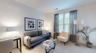 This single-floor residence is thoughtfully designed with a modern aesthetic and functional open-concept layout. The living area serves as the heart of the space, featuring neutral-toned carpet, large windows dressed with sheer curtains that invite abundant natural light and offer views of greenery outside. The room comfortably accommodates a full seating arrangement centered around a glass oval coffee table.

The kitchen is a standout feature, showcasing rich dark espresso cabinetry paired with granite countertops and a central island with bar seating. Stainless steel appliances, including a gas range, built-in microwave, and refrigerator, complement the warm hardwood flooring. Pendant lighting and a track light system illuminate the workspace effectively.

The bedroom is a calm retreat with neutral walls, plush carpet, and dual windows overlooking outdoor greenery. It provides ample space for a full bedroom suite and benefits from natural light throughout the day.

The bathroom maintains the home's clean design language with dark vanity cabinetry, a white countertop, large-format stone-look tile flooring, and a full tub-shower combination with white tile surround. Throughout the residence, a consistent palette of whites, grays, and warm wood tones creates a cohesive, contemporary ambiance.