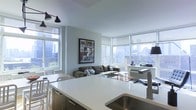 This single-floor space showcases a thoughtfully designed open-concept layout that integrates the kitchen and living areas into one cohesive environment. The kitchen features sleek, dark cabinetry paired with a bright white countertop island, providing both functional workspace and casual seating. A stainless steel sink adds a clean, contemporary finish to the space.

Large floor-to-ceiling windows wrap around the living area, flooding the interior with abundant natural light while framing impressive panoramic city skyline views. The windows are fitted with roller shades, offering flexible light control. The living area is anchored by a comfortable sectional sofa in a neutral tone, complemented by a framed urban art piece that adds personality to the light gray walls.

Industrial-style pendant lighting above the kitchen island enhances the modern aesthetic, while the light hardwood flooring ties the open space together seamlessly. The overall ambiance is contemporary and airy, with a clean, minimalist design philosophy that lets the striking cityscape serve as the primary visual focal point throughout the space.