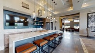 Located at 9295 One Deerfield Pl. in Mason, Ohio, this single-floor community clubhouse offers a sophisticated and thoughtfully designed gathering space. The entrance greets visitors with large-format slate tile flooring, soaring ceilings, and a dramatic oversized clock above glass-paneled double doors, immediately establishing the upscale tone of the space.

The kitchen and bar area serves as a central hub, featuring an expansive marble-topped island with leather-upholstered barstools, teal subway tile backsplash, open wood shelving, stainless steel appliances, and a suite of eclectic pendant lights ranging from smoked glass to crystal. A dedicated coffee and beverage station adds everyday convenience.

Adjacent to the kitchen, a cozy lounge area is anchored by a sleek linear fireplace set against a stone-clad accent wall, paired with rich cognac leather sectionals, textured area rugs, and floor-to-ceiling windows that draw in generous views of the surrounding landscape. Wall sconces and recessed lighting create a warm, inviting atmosphere.

The dining area is equally impressive, showcasing double-height ceilings with clerestory windows, herringbone-patterned wood-look tile flooring, multiple dining tables with upholstered chairs, and a striking large-scale botanical wall mural. A mounted display screen and abundant outdoor views complete the space, making it ideal for both casual gatherings and organized events.