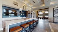 Located at 9295 One Deerfield Pl. in Mason, Ohio, this single-floor community clubhouse offers a sophisticated and thoughtfully designed gathering space. The entrance greets visitors with large-format slate tile flooring, soaring ceilings, and a dramatic oversized clock above glass-paneled double doors, immediately establishing the upscale tone of the space.

The kitchen and bar area serves as a central hub, featuring an expansive marble-topped island with leather-upholstered barstools, teal subway tile backsplash, open wood shelving, stainless steel appliances, and a suite of eclectic pendant lights ranging from smoked glass to crystal. A dedicated coffee and beverage station adds everyday convenience.

Adjacent to the kitchen, a cozy lounge area is anchored by a sleek linear fireplace set against a stone-clad accent wall, paired with rich cognac leather sectionals, textured area rugs, and floor-to-ceiling windows that draw in generous views of the surrounding landscape. Wall sconces and recessed lighting create a warm, inviting atmosphere.

The dining area is equally impressive, showcasing double-height ceilings with clerestory windows, herringbone-patterned wood-look tile flooring, multiple dining tables with upholstered chairs, and a striking large-scale botanical wall mural. A mounted display screen and abundant outdoor views complete the space, making it ideal for both casual gatherings and organized events.