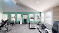 This fitness center offers a comprehensive exercise environment designed to support a wide range of workout routines. The space is defined by striking vaulted ceilings that rise dramatically, painted in a bold teal that transitions into light gray walls, creating an energizing and visually dynamic atmosphere. Large windows fitted with blinds line the walls, allowing ample natural light to fill the room throughout the day.

The main exercise area is outfitted with a variety of cardio machines, including treadmills, elliptical trainers, and a stair stepper, arranged to maximize floor space and ease of movement. A rack of free weights and a water station provide added convenience for users. Strength training equipment, including a multi-function cable and press machine, is positioned in a separate area alongside a dedicated stretching zone complete with resistance bands, a stability ball, foam rollers, and a wall-mounted mirror.

A wood-paneled accent wall features an inspirational quote — "On good days, work out. On bad days, work out harder" — adding a motivational focal point to the space. A ceiling fan overhead aids in air circulation. Locker storage units near the entrance offer secure storage options. The floor is covered in durable, textured carpet tiles, and modern recessed lighting complements the natural light, ensuring the space remains bright and functional at all hours.