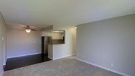 This single-floor space offers an open-concept layout that seamlessly connects the living area to the kitchen. The living area is finished with plush, neutral-toned carpeting and painted in soft gray tones, creating a calm and inviting atmosphere. A ceiling fan is visible, contributing to both comfort and functionality. The textured ceiling adds a subtle design detail consistent throughout the space.

The kitchen is partially separated by a raised breakfast bar with a dark countertop, providing a natural division between the cooking and living zones while maintaining an open feel. White cabinetry lines the kitchen walls, offering ample storage, while a stainless steel refrigerator adds a modern touch. The kitchen features hard flooring, contrasting with the carpeted living area and providing a practical surface for the cooking space.

The pass-through design of the kitchen enhances sightlines across the unit, making the overall space feel larger and more connected. Clean white baseboards frame each room, and the neutral wall colors throughout create a cohesive, move-in-ready aesthetic suitable for a variety of interior styles.