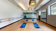Located on S Alton St in Denver, CO, this Tava Waters fitness studio offers a thoughtfully designed, single-floor group exercise space ideal for barre, yoga, and mat-based classes. The studio features warm hardwood flooring throughout, providing both aesthetic appeal and a functional surface for a variety of workouts. Along one full wall, a double-tiered wooden ballet barre is mounted at varying heights to accommodate participants of all statures, a hallmark of a well-equipped barre studio. Large, floor-to-ceiling mirrors are positioned on multiple walls, allowing participants to monitor their form from multiple angles, while also amplifying the sense of openness within the space. Natural light filters in through several windows, offering views of the surrounding tree-lined landscape and contributing to an energizing atmosphere. The studio is equipped with branded Tava Waters yoga mats and foam blocks, ready for class use. A wall-mounted flat-screen display supports instructor-led or on-demand fitness programming. Overhead, distinctive woven rattan pendant light fixtures add a warm, boutique-studio ambiance that contrasts elegantly with the clean white and gray walls. The entry is framed by frosted glass panels, ensuring privacy while maintaining a modern aesthetic. Ample room allows for comfortable, simultaneous participation by multiple guests.