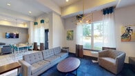 Located at 1028 E St Louis St in Springfield, Missouri, this single-floor community clubhouse offers a thoughtfully designed, contemporary gathering space that blends comfort, recreation, and productivity. The open main area features large-format tile flooring and soaring ceilings, creating a bright and spacious atmosphere. Bold pendant lighting with gold-interior shades anchors the central communal dining area, furnished with long dark tables and teal chairs that reflect the space's vibrant color palette.

Adjacent to the dining zone, a well-appointed lounge area showcases plush tufted sofas, armchairs, and a rich blue carpet beneath a striking geometric brass chandelier. Oversized windows throughout invite abundant natural light and provide pleasant outdoor views. Colorful artwork adds personality and energy to the walls.

The game room area includes a regulation pool table, an arcade cabinet, and a flat-screen TV mounted against a decorative feature wall with patterned wallcovering — ideal for leisure and socializing. A dedicated business center, enclosed with glass sliding doors, provides a quiet workspace equipped with desks, ergonomic seating, and office equipment. A small seating area just outside the business center offers additional convenience for visitors. The overall design is modern, energetic, and well-suited for community engagement.
