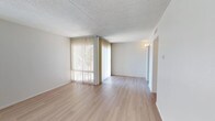 This single-floor space offers a clean and functional layout designed to maximize comfort and usability. The main living area is immediately welcoming, featuring light-toned hardwood flooring that runs throughout and lends a warm, cohesive feel to the space. A prominent wall of floor-to-ceiling vertical blinds allows for generous natural light while offering privacy control, creating a bright and airy atmosphere. The textured ceiling adds a classic architectural detail, while the neutral white walls provide a versatile backdrop that complements a wide range of furnishing styles. A white-painted brick accent wall adds a subtle textural element and visual interest to the room. A wall-mounted light fixture near the hallway provides additional ambient lighting, guiding the transition into the rest of the space. The open layout promotes a sense of spaciousness, making the area feel expansive and easy to arrange. An HVAC vent is integrated neatly into the upper wall, ensuring year-round climate comfort. Overall, the space presents a well-maintained, light-filled interior that balances simplicity with functional design details.