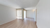 This single-floor space offers a clean and functional layout designed to maximize comfort and usability. The main living area is immediately welcoming, featuring light-toned hardwood flooring that runs throughout and lends a warm, cohesive feel to the space. A prominent wall of floor-to-ceiling vertical blinds allows for generous natural light while offering privacy control, creating a bright and airy atmosphere. The textured ceiling adds a classic architectural detail, while the neutral white walls provide a versatile backdrop that complements a wide range of furnishing styles. A white-painted brick accent wall adds a subtle textural element and visual interest to the room. A wall-mounted light fixture near the hallway provides additional ambient lighting, guiding the transition into the rest of the space. The open layout promotes a sense of spaciousness, making the area feel expansive and easy to arrange. An HVAC vent is integrated neatly into the upper wall, ensuring year-round climate comfort. Overall, the space presents a well-maintained, light-filled interior that balances simplicity with functional design details.