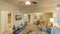 This single-floor residence offers a thoughtfully designed open-concept layout that seamlessly connects the living, dining, and kitchen areas. The living room is a comfortable and inviting space, anchored by plush carpeting and accented with classic crown molding that runs along the ceiling perimeter. A ceiling fan with an integrated light fixture adds both functionality and a traditional charm to the room. A standout architectural feature is the graceful arched passageway that connects the living area to the dining space, lending the interior a refined, cohesive character. The dining area accommodates a round table with upholstered chairs, complemented by a pendant light overhead and framed artwork on the walls, creating an intimate setting for meals. Beyond the arch, the kitchen is accessible and conveniently positioned near a counter-height bar seating area. Throughout the space, neutral warm tones on the walls and flooring create a calm, welcoming ambiance. The layout promotes easy flow between rooms, making the residence both practical and aesthetically pleasing. Overall, this single-floor home combines classic architectural details with comfortable, functional living spaces.