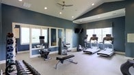This fitness center offers a thoughtfully designed workout environment that combines functionality with an inviting aesthetic. The space is characterized by vaulted ceilings, recessed lighting, and a ceiling fan that keep the area bright and comfortable during exercise. Deep teal-blue walls provide a bold, energizing backdrop, while large windows along multiple walls allow natural light to fill the room and offer pleasant outdoor views.

The facility is well-appointed with a variety of cardio and strength training equipment. Multiple treadmills are positioned near the windows, allowing users to enjoy natural light during their workouts. A stationary bike, cable strength machine, and a weight bench are also available, catering to a range of fitness routines. A set of free weights is conveniently arranged for easy access. A wall-mounted flat-screen television adds an entertainment option for those looking to stay engaged during longer sessions.

A large wall mirror enhances the sense of space and provides users with a functional view for form-checking during strength exercises. The neutral-toned carpet and clean white trim complement the rich wall color, creating a polished and cohesive interior. Overall, this fitness center strikes a balance between practical equipment offerings and an attractive, well-maintained environment.