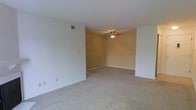 This single-floor space features an open living area designed with simplicity and functionality in mind. The main living space is finished with wall-to-wall neutral-toned carpeting, providing a soft, comfortable underfoot feel that extends throughout the room. The walls are painted in a warm, light beige tone, contributing to an inviting and cohesive atmosphere.

A ceiling fan with an integrated light fixture is centrally mounted, offering both ambient lighting and air circulation. Ventilation is well-considered, with visible HVAC registers positioned along the walls and near the baseboard, ensuring efficient climate control throughout the space. Baseboard trim lines the perimeter of the room, adding a clean, finished look to the flooring transition.

The layout is open and unobstructed, allowing for flexible furniture arrangement to suit a variety of needs. The room transitions naturally into an adjacent area, suggesting a connected, flowing floor plan. Overall, the space presents a clean, move-in ready environment with practical features suited for comfortable everyday living.
