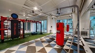 Located at 20 Water Grant St in Yonkers, NY, this expansive fitness center delivers a comprehensive workout experience across a generous single-floor layout. The space is thoughtfully designed with a bold, energetic aesthetic, featuring eye-catching geometric-patterned flooring in shades of blue, gray, and white that covers the main exercise areas, while vibrant artificial turf defines dedicated functional training zones.

The strength and conditioning area is well-appointed with a cable machine, weight benches, a power rack with plate-loaded barbells, dumbbells, and medicine balls, all set against a striking accent wall of bold teal, navy, and green vertical stripes. A wall-mounted flat-screen TV adds convenience for workout entertainment. Full-length mirrors line portions of the walls, enhancing the sense of space and supporting form-focused training.

The functional training zone features a heavy-duty rig, red hanging punching bags, agility ladder markings, and a large tire — ideal for high-intensity and athletic conditioning workouts. Track lighting and exposed ductwork on the high ceilings contribute to the facility's modern, industrial-inspired character.

Large glass sliding doors open directly to an outdoor courtyard and pool area, allowing natural light to flood the space while providing seamless indoor-outdoor connectivity. The overall atmosphere is vibrant, motivating, and professionally equipped.