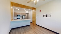 Located at 3212 Jaffa Garden Way in Rapid City, South Dakota, this single-floor unit offers a clean, contemporary design with consistent wood-look flooring and a neutral color palette throughout. The open-concept kitchen is a standout feature, centered around a bold two-tone island with a white countertop and navy blue base. Warm natural wood paneling serves as a striking architectural accent, framing the kitchen and lending visual warmth to the space. The kitchen is fully equipped with stainless steel appliances, including an electric range with a built-in microwave overhead, complemented by light wood cabinetry with modern hardware and white countertops. Pendant lighting above the island adds a refined finishing touch. The adjacent living area is open and well-lit, benefiting from a large window that invites natural light and outdoor views. A ceiling fan, wall-mounted TV bracket, and baseboard heating complete the room. The hallway leads to a bathroom featuring a walk-in shower with white subway tile surround and built-in shelving, finished with brushed nickel fixtures. An in-unit washer is also conveniently accessible from the hallway, adding everyday practicality to this thoughtfully designed space.