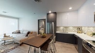 This contemporary single-floor residence in Los Angeles, CA (91325) offers a well-designed open-concept layout that seamlessly connects the kitchen, dining, and living areas. The kitchen is outfitted with stainless steel appliances including a French door refrigerator, range, and over-the-range microwave, complemented by two-tone cabinetry in dark charcoal and crisp white, white quartz countertops, and a distinctive hexagonal mosaic tile backsplash. Light wood-look flooring runs throughout the main living areas, adding warmth to the modern aesthetic. The dining area features a wooden table with contemporary seating, positioned centrally between the kitchen and living room. The living room offers a large window with a roller shade, providing controlled natural light. The primary bedroom features plush carpeting, a vibrant teal accent wall, a walk-in closet with built-in shelving, and a dedicated en-suite bathroom with an LED backlit vanity mirror and dark cabinetry. The second bedroom is generously sized with carpeted floors, natural light from a window, and a built-in desk area. Two bathrooms mirror each other in design, both featuring LED-lit vanity mirrors, dark-toned under-sink cabinetry, and white quartz countertops, with one offering a shower-tub combination.