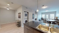This well-appointed single-floor residence offers a thoughtfully designed open-concept layout that seamlessly connects the kitchen, dining, and living areas. The contemporary kitchen serves as the heart of the unit, anchored by a generous island with a dark granite-style countertop, an undermount sink, and bar seating for two. Pendant lighting hangs above the island, adding warmth to the space. The kitchen is fully equipped with stainless steel appliances, including a French door refrigerator, electric range, and microwave, complemented by dark wood cabinetry that extends to the upper walls. Light hardwood-style flooring runs throughout the main living areas, creating a clean and cohesive aesthetic. A dedicated in-unit laundry closet houses a stacked washer and dryer, offering added convenience. The living room is bright and inviting, featuring a set of French doors that open to a private balcony with views of the surrounding neighborhood, allowing for ample natural light. The bedroom is carpeted for comfort and includes a large window with slatted blinds that fill the room with soft, filtered light. A full bathroom with a shower/tub combination and neutral finishes completes the layout. The overall design palette of soft grays, warm wood tones, and clean white accents creates a modern yet welcoming ambiance throughout.