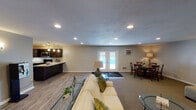 This single-floor space is designed around an open-concept layout that seamlessly connects the kitchen, living area, dining nook, and a separate lounge zone. The kitchen is a standout feature, outfitted with rich dark espresso cabinetry, light granite countertops, stainless steel appliances including a range, microwave, dishwasher, and refrigerator, and pendant lighting that adds a polished, modern touch. A generous island or peninsula extends the counter space and reinforces the kitchen's functionality.

The main living area transitions smoothly from wood-look hard flooring near the kitchen to carpeted zones, creating defined areas for relaxation and entertaining. French doors along one wall introduce natural light and provide a view of an outdoor area beyond. A round dining table with cross-back chairs occupies a cozy corner near a side door, accompanied by a wall-mounted air conditioning unit and floor vent for climate control.

A secondary lounge area features plush patterned armchairs, ottomans, and a glass-top side table, illuminated by recessed ceiling lights and wall-mounted accent fixtures. Neutral gray walls throughout unify the space, while recessed lighting provides consistent, even illumination across all areas, contributing to a clean and welcoming ambiance.
