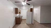 This single-floor unit features an open-concept layout that connects the kitchen and living area into one cohesive space. The kitchen is equipped with dark wood cabinetry, both upper and lower, providing ample storage. A double-basin sink sits beneath the cabinets along the countertop, which features a dark laminate surface. A full-size white refrigerator stands at the end of the kitchen run, with additional upper cabinetry mounted above it for extra storage. The kitchen flooring consists of a patterned vinyl tile, offering a practical and easy-to-clean surface. The living area transitions seamlessly from the kitchen, featuring neutral carpet that extends throughout. A ceiling fan with a light fixture is centrally positioned, serving both the kitchen and adjacent living space. The walls are painted in a clean, neutral white, and the textured ceiling adds a subtle visual detail. A hallway extends from the living area, suggesting additional rooms beyond the main open space. The overall layout is functional and straightforward, making efficient use of the available floor plan.